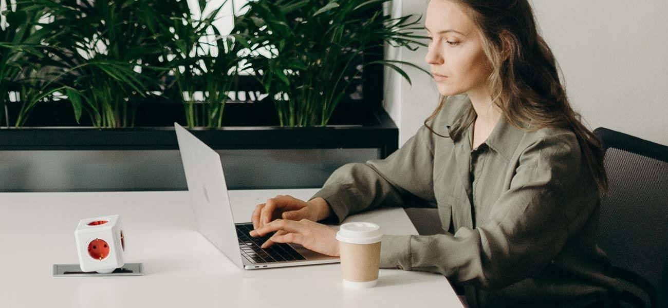 Woman working on a laptop at a desk with a coffee cup and a power outlet, surrounded by greenery, illustrating email marketing strategies and remote work productivity.