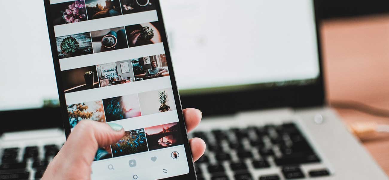 Hand holding smartphone displaying social media feed with images, alongside a laptop, illustrating social media marketing strategies for engaging customers.
