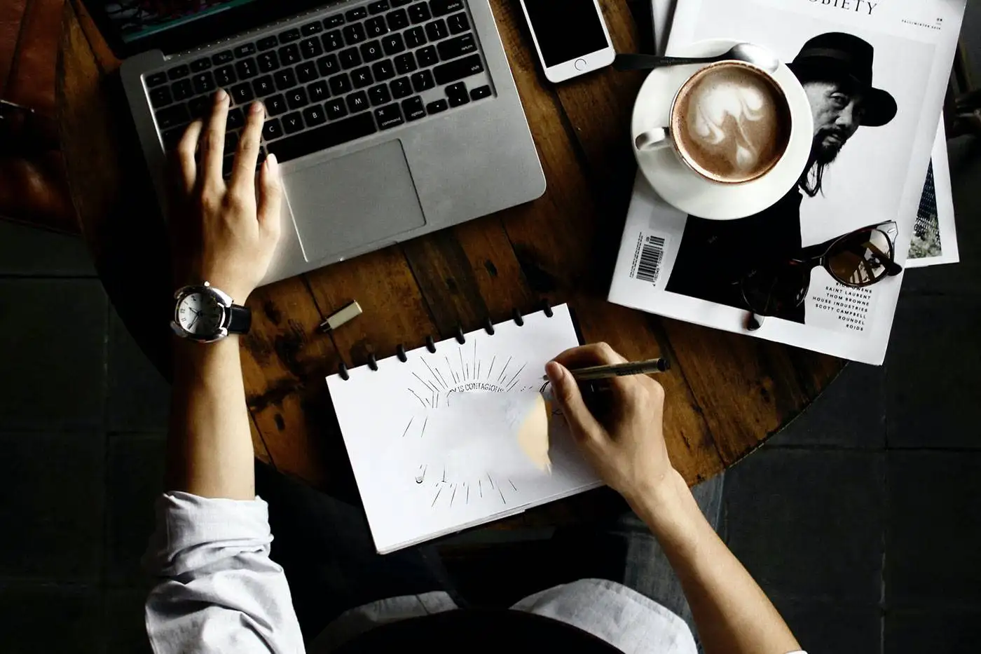 Person writing in a notebook next to a laptop and a cup of coffee, illustrating the creative process in a workspace.