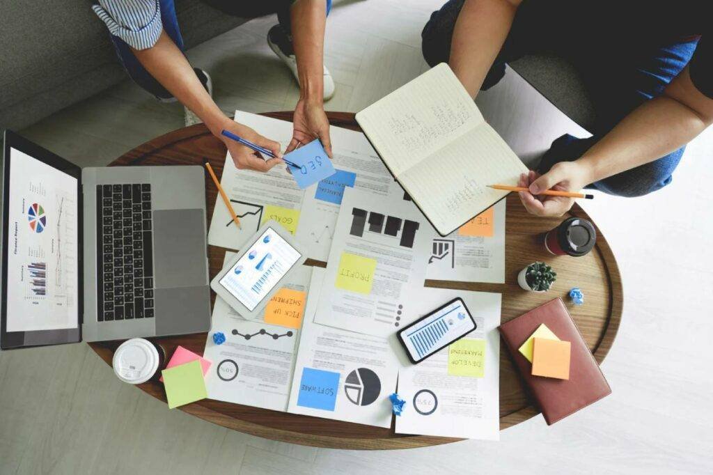 Collaborative workspace with laptop displaying graphs, documents, sticky notes, and two individuals analyzing data for sales automation solutions.