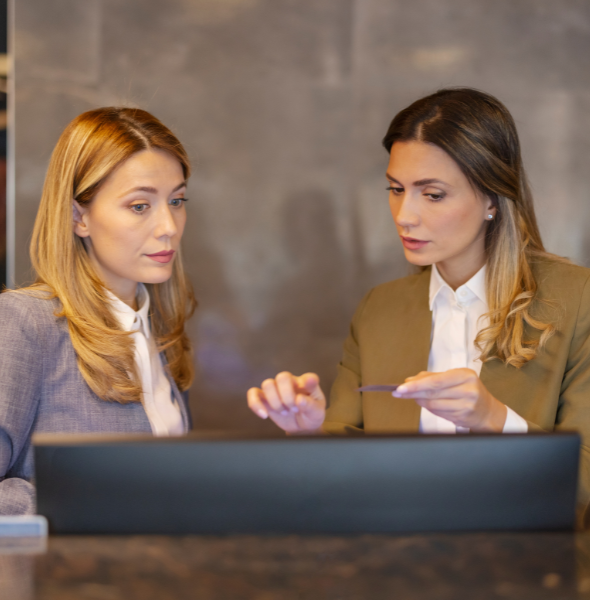 Two professional women discussing information at a computer, representing collaboration for beverage catering inquiries and bookings.
