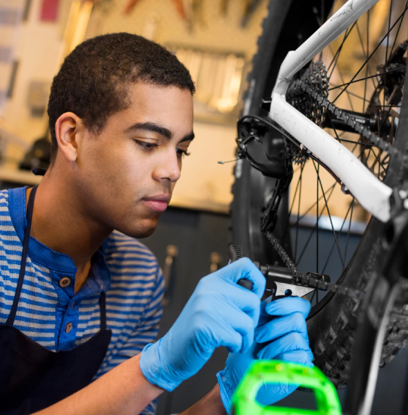 Young mechanic repairing a bicycle, wearing blue gloves, focused on adjusting the bike's components, with tools and a workbench in the background, illustrating hands-on service in a bicycle repair shop.